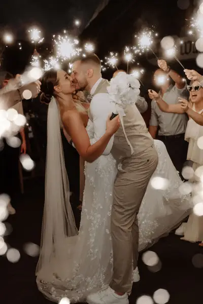 Hochzeitspaar küsst sich bei Nacht unter Lichtern, umgeben von Gästen mit Wunderkerzen.